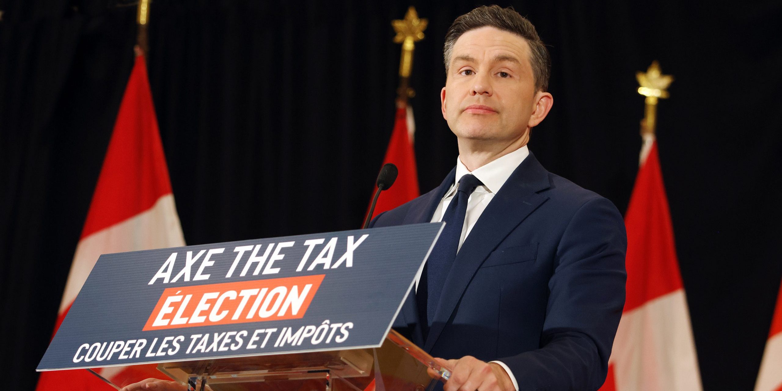 Pierre Poilievre stands behind a clear podium, on which a "Axe the Tax" election photo is displayed. He stands in front of a row of Canadian flags.